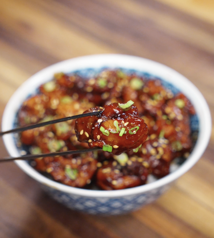A close-up of glossy sesame chicken held between chopsticks, showing the sticky sauce, sesame seeds, and scallions.
