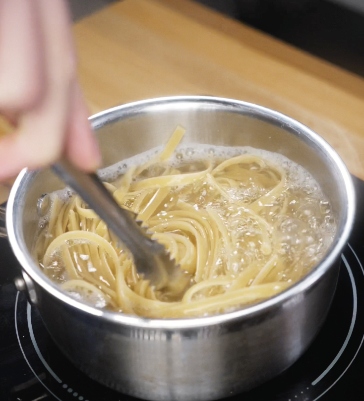 Fettuccini noodles are stirred in a pot of boiling water with tongs until nearly al dente, just before draining.