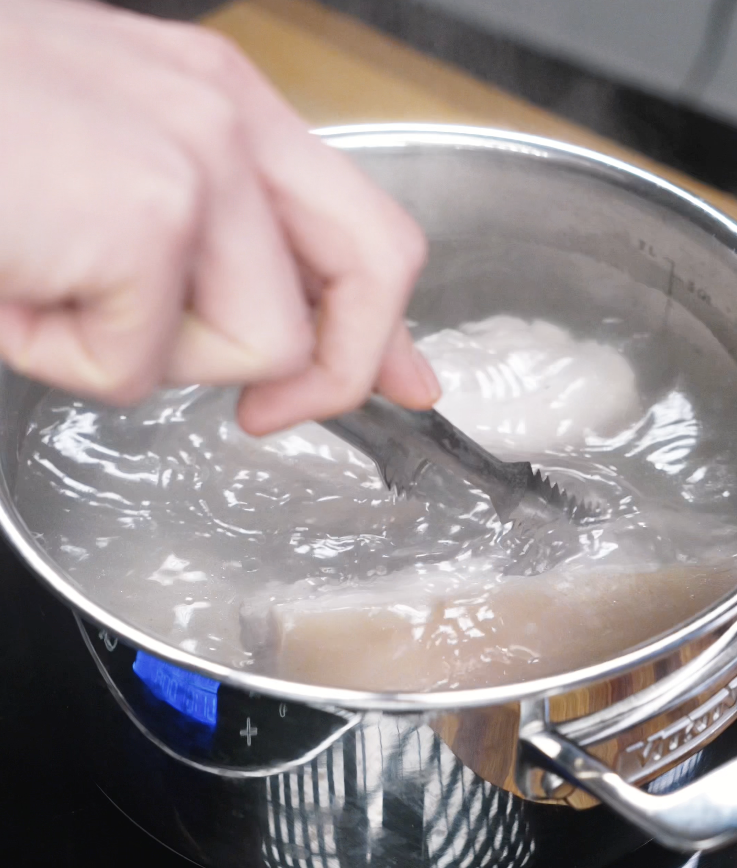 Pork belly is being blanched in boiling water using tongs to remove impurities before air frying.