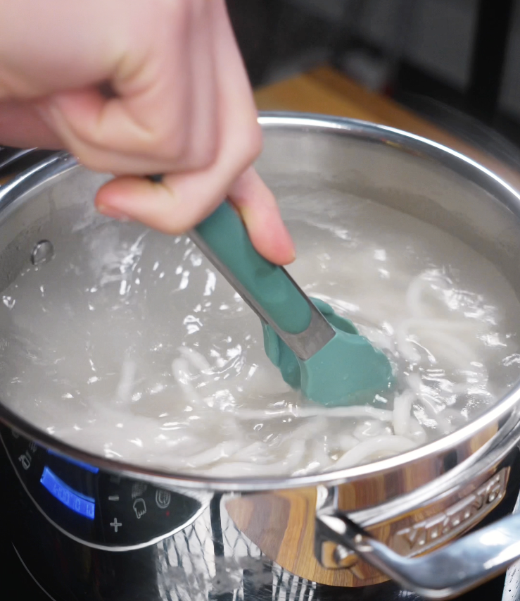 Thick udon noodles are stirred in boiling water with tongs until tender and chewy.