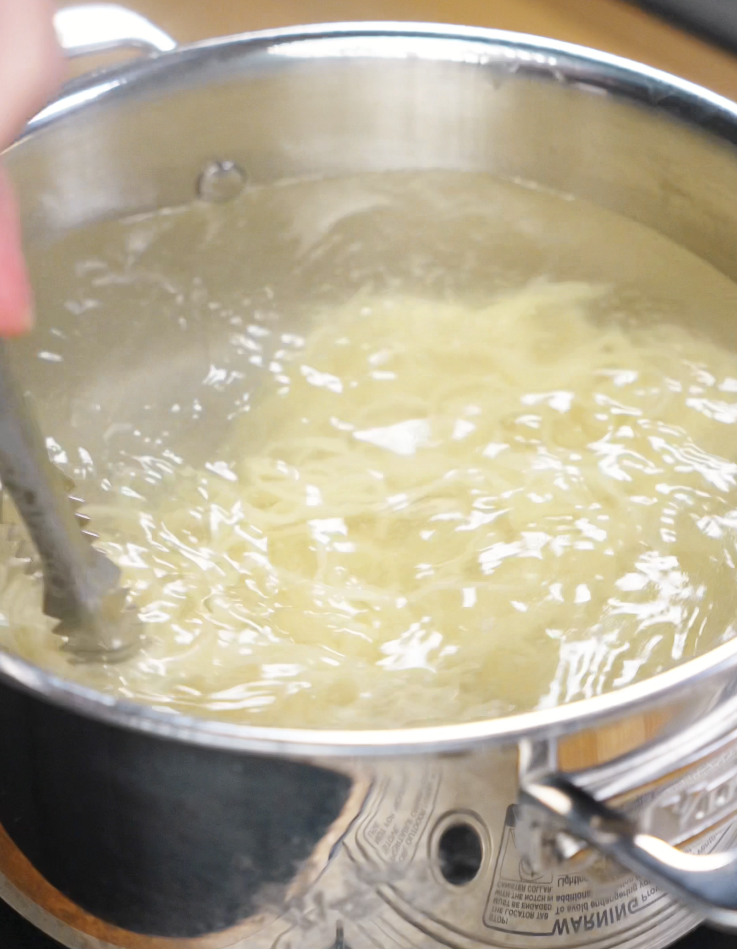 Chow mein noodles swirl in a pot of boiling water, being stirred gently with metal tongs.