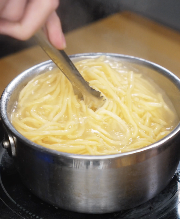 Fresh noodles are cooked until al dente in a pot of boiling water, stirred gently with metal tongs.