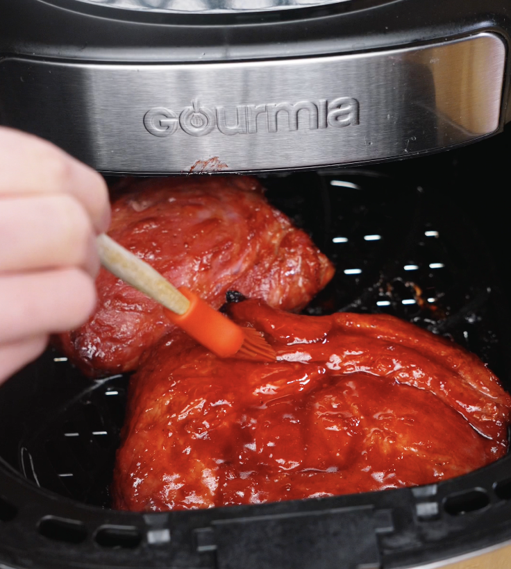A silicone brush applying a layer of char siu glaze onto pork in the air fryer halfway through cooking.
