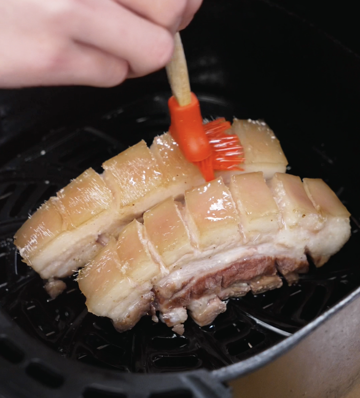 The skin of partially cooked pork belly is brushed with neutral oil inside the air fryer basket.