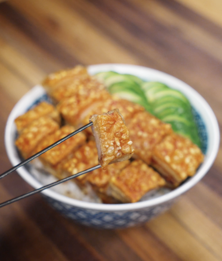 A crispy cube of pork belly held up with chopsticks, highlighting the golden skin and juicy layers of meat.