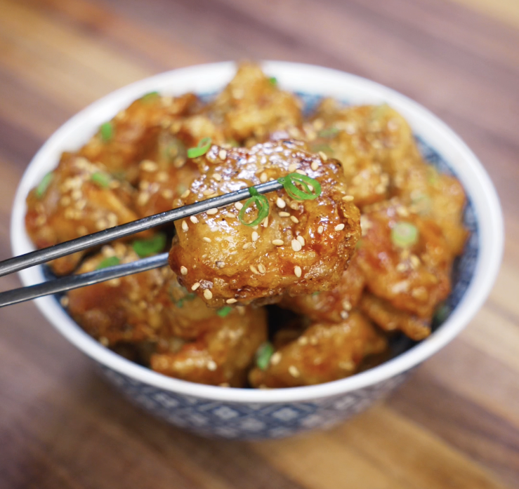 Chopsticks holding a glistening piece of honey butter chicken over a bowl, showing the sesame-studded glaze and juicy texture.