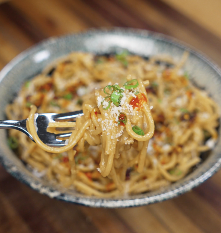 A fork lifts a twirl of creamy miso butter pasta topped with chili crisp, scallions, and a sprinkle of cheese, showcasing the silky sauce coating.