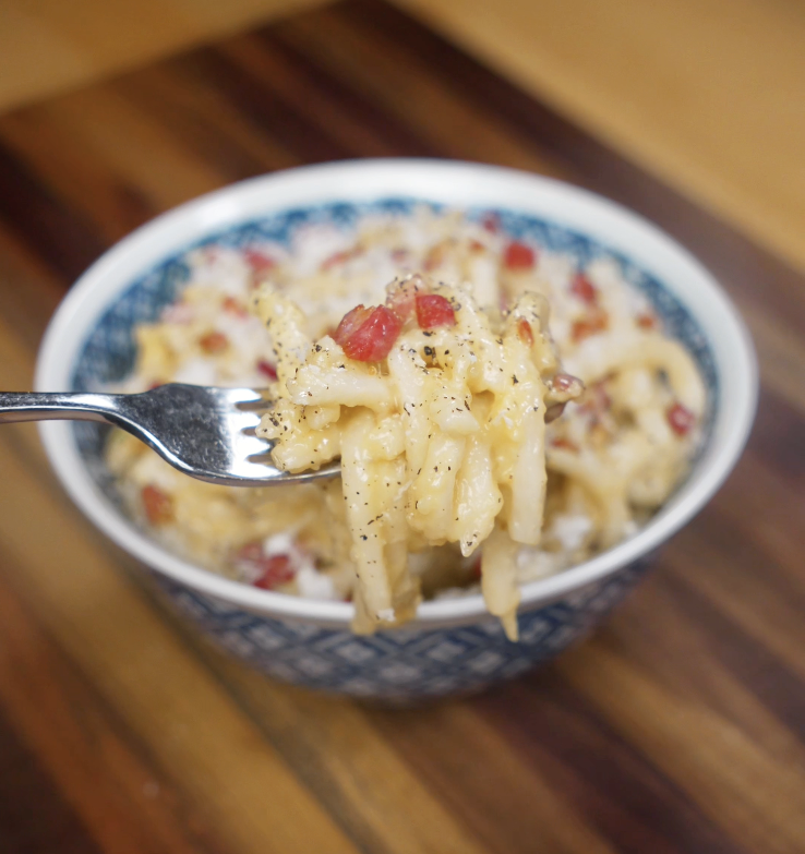 A fork lifts a bite of creamy carbonara udon showing glossy noodles coated in sauce with bits of pancetta and black pepper.
