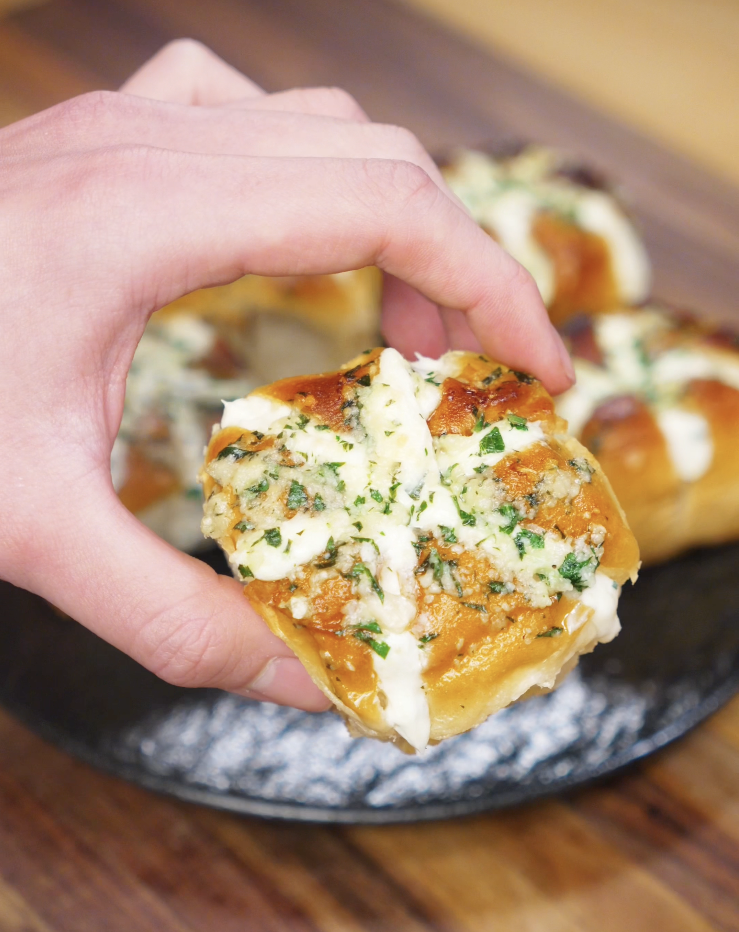 A hand holds up a finished Korean Garlic Cheese Bread roll, showing the golden top, cheesy filling, and parsley garlic glaze.