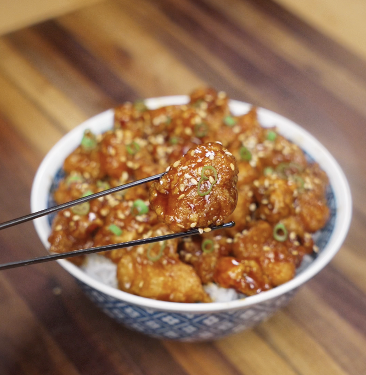 A piece of sticky glazed General Tso’s Chicken lifted with chopsticks over the rice bowl.