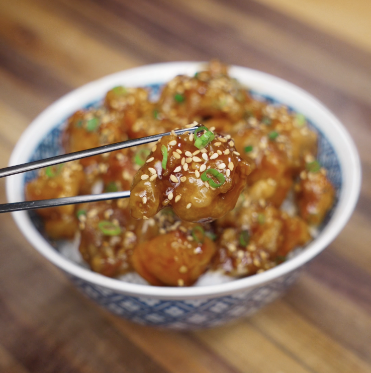 A glossy, golden piece of Korean fried chicken is lifted from the bowl with chopsticks, showing the sticky glaze.