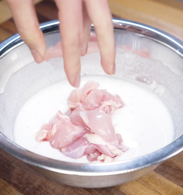 Raw chicken thigh chunks being dropped into a bowl of white batter for a light, crispy coating.