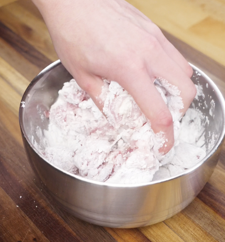 Chicken chunks being coated in a flour and cornstarch mixture by hand in a metal mixing bowl.