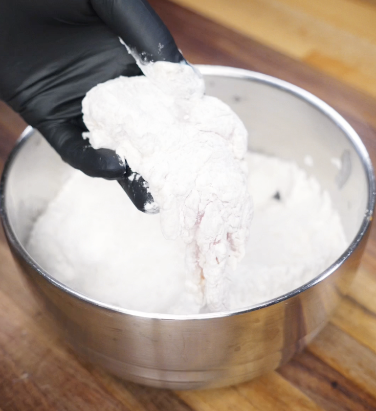 A gloved hand lifts a marinated chicken tender coated in flour mixture, making sure every crevice is covered for maximum crunch.