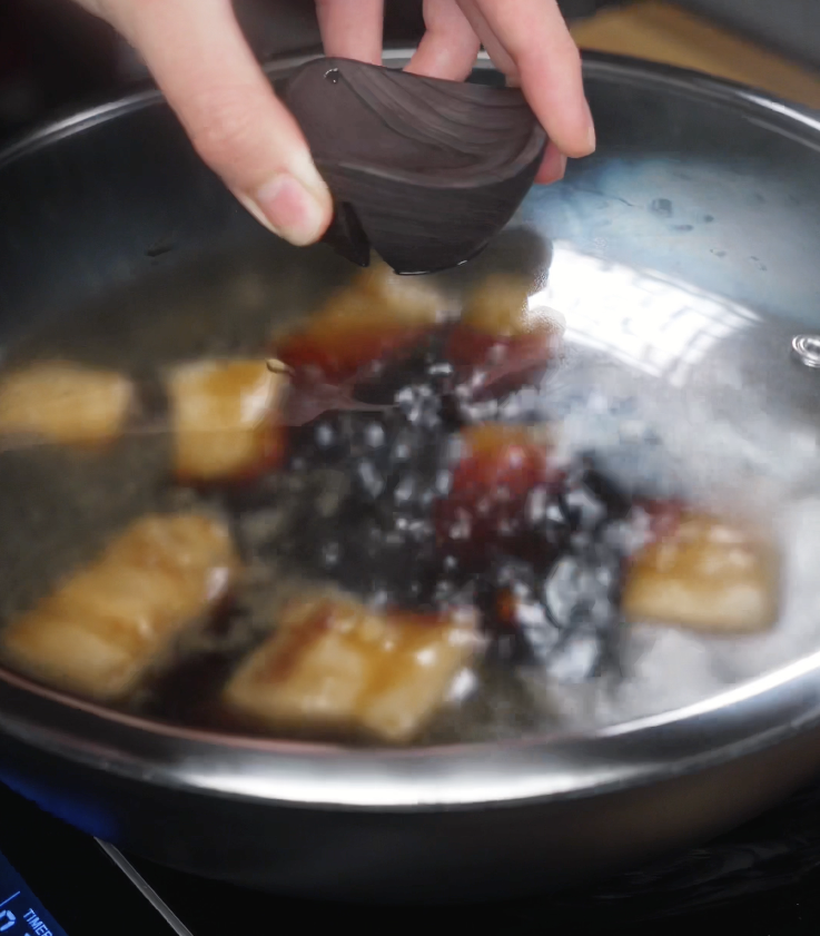 A hand places the lid onto the pot to cover simmering pork belly in a rich soy-based braising liquid, allowing it to cook low and slow.