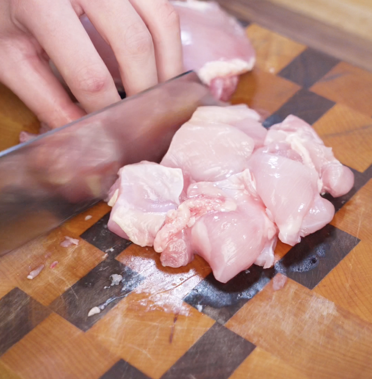 Raw boneless chicken thighs being cut into chunks on a wooden cutting board in preparation for frying.