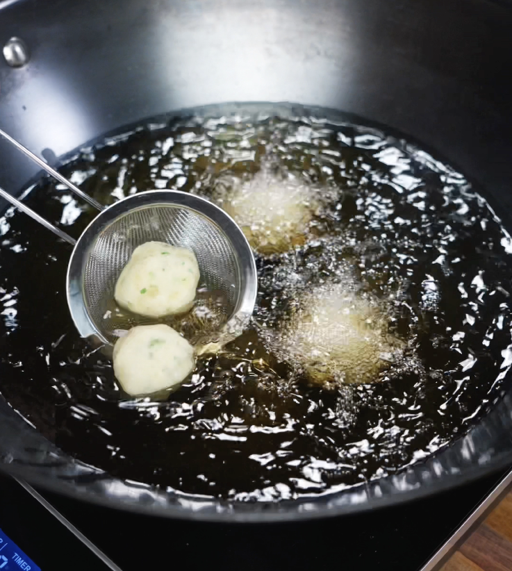 Potato balls are gently lowered into hot oil using a fine mesh strainer, beginning their transformation into crispy bites.