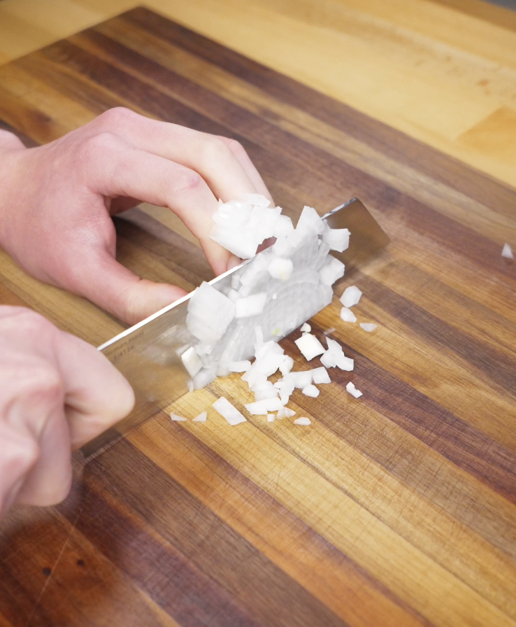 A hand finely dices a white onion on a wooden cutting board with a chef’s knife.