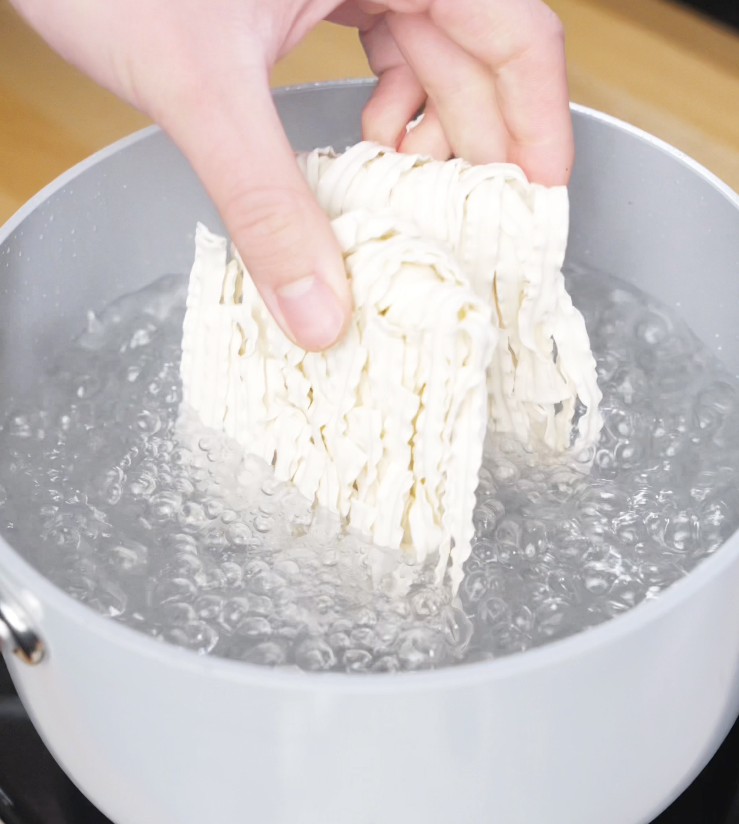 A hand dropping a block of instant noodles into a pot of rapidly boiling water, marking the start of the noodle prep for chili garlic butter noodles.