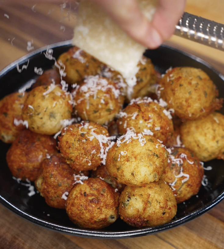 A hand grates fresh parmesan over a platter of golden brown potato balls, adding a finishing touch of salty richness and visual appeal.