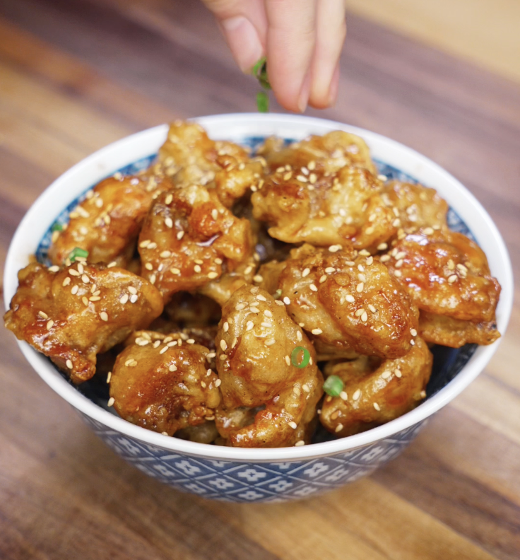 A hand sprinkling fresh scallions over a bowl of glossy honey butter chicken with sesame seeds.
