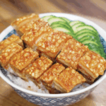 A bowl of crispy pork belly over steamed rice, garnished with fresh cucumber slices on the side.