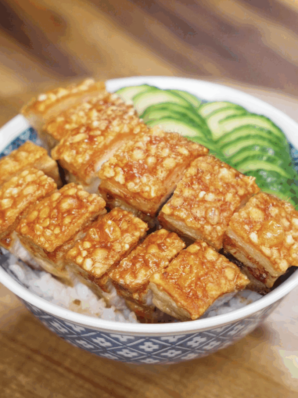A bowl of crispy pork belly over steamed rice, garnished with fresh cucumber slices on the side.