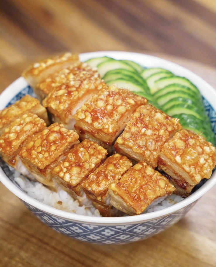 A bowl of crispy pork belly over steamed rice, garnished with fresh cucumber slices on the side.