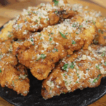 A pile of garlic parmesan chicken tenders sits on a black plate, perfectly golden and topped with buttery garlic, parmesan, and fresh parsley.