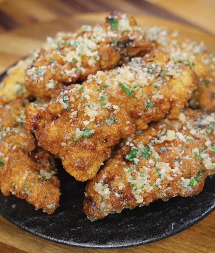 A pile of garlic parmesan chicken tenders sits on a black plate, perfectly golden and topped with buttery garlic, parmesan, and fresh parsley.