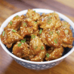 A bowl filled with golden, crispy honey butter chicken garnished with sesame seeds and scallions, ready to serve.