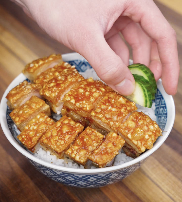 A bowl of crispy pork belly over steamed rice, garnished with fresh cucumber slices on the side.