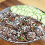 A generous bowl of garlic steak bites over white rice, topped with parsley and parmesan, and fanned cucumber slices on the side.