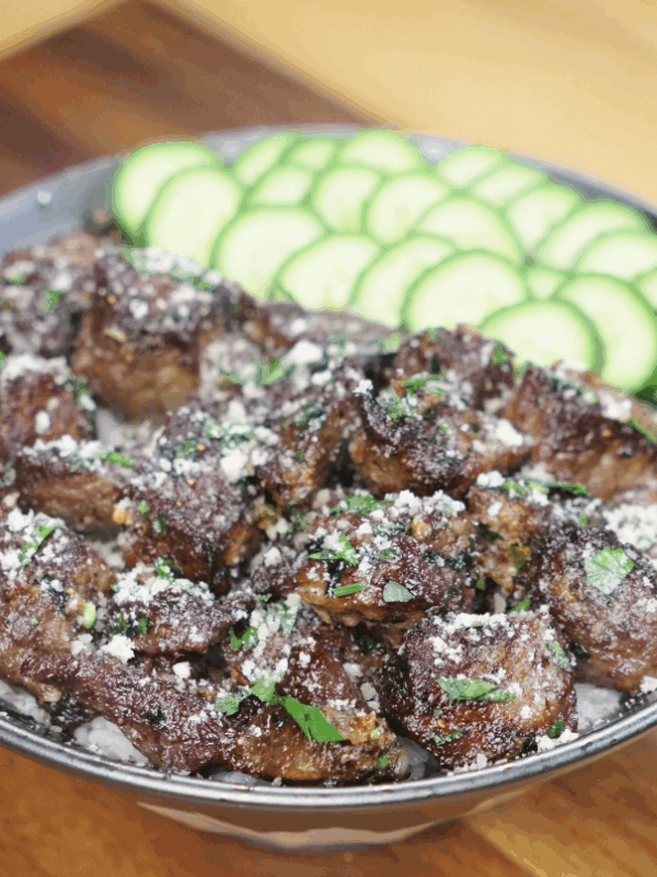 A generous bowl of garlic steak bites over white rice, topped with parsley and parmesan, and fanned cucumber slices on the side.
