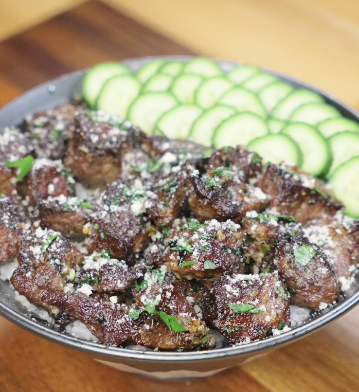 A generous bowl of garlic steak bites over white rice, topped with parsley and parmesan, and fanned cucumber slices on the side.