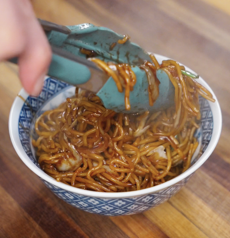Saucy chow mein noodles are lifted from the pan and served steaming hot into a patterned blue bowl.