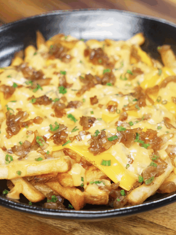 A skillet filled with golden fries topped with gooey American cheese, caramelized onions, and a creamy gochujang animal sauce, finished with chives.