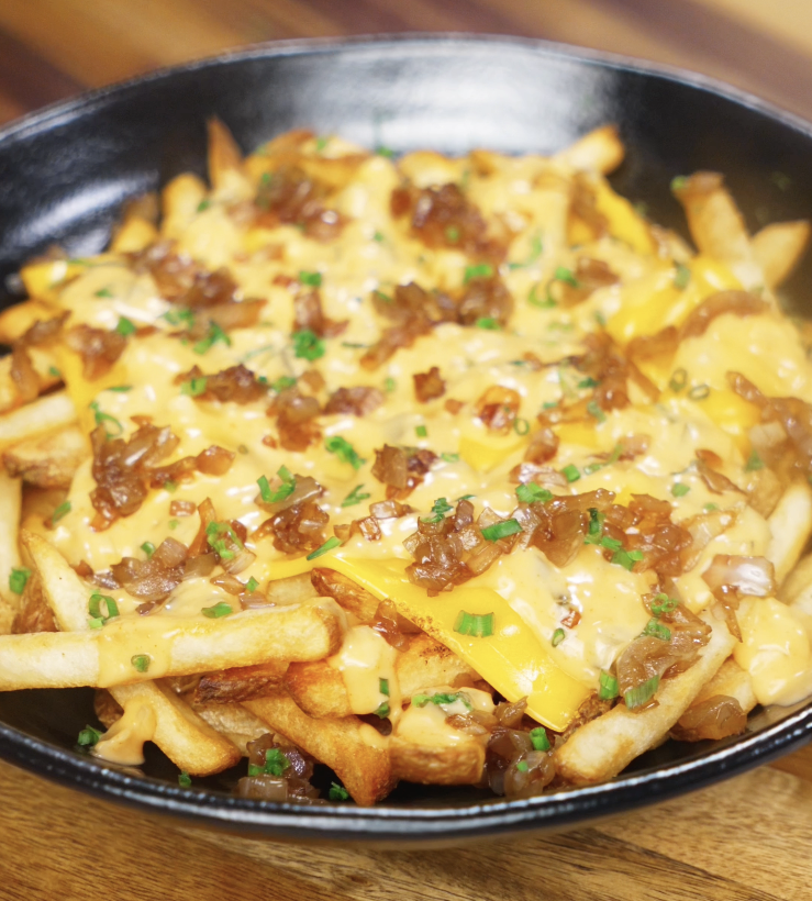 A skillet filled with golden fries topped with gooey American cheese, caramelized onions, and a creamy gochujang animal sauce, finished with chives.