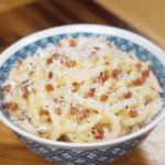 A bowl of creamy carbonara udon topped with pancetta, parmesan, and black pepper sits ready to serve.