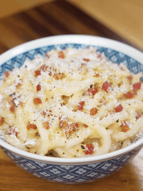 A bowl of creamy carbonara udon topped with pancetta, parmesan, and black pepper sits ready to serve.
