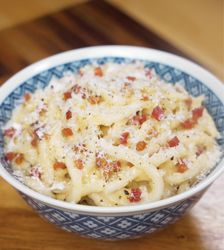 A bowl of creamy carbonara udon topped with pancetta, parmesan, and black pepper sits ready to serve.