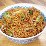 A bowl of chow mein with glossy noodles, stir-fried onions, scallions, and cabbage, ready to eat.