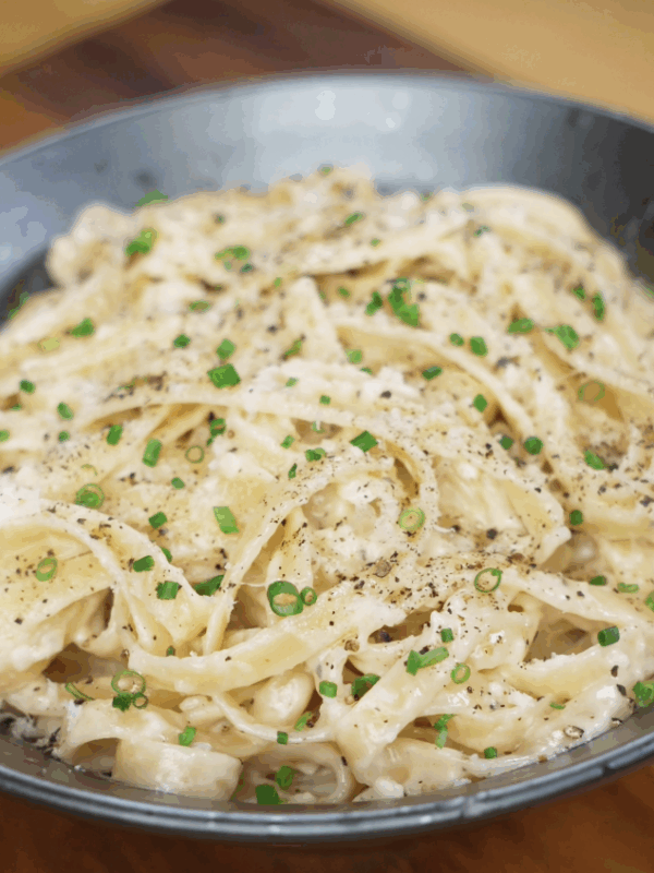 A bowl of creamy garlic fettuccini Alfredo is topped with freshly cracked black pepper and finely chopped chives for a rich, aromatic finish.