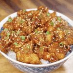 A bowl of General Tso’s Chicken served over white rice, topped with sesame seeds and sliced scallions.