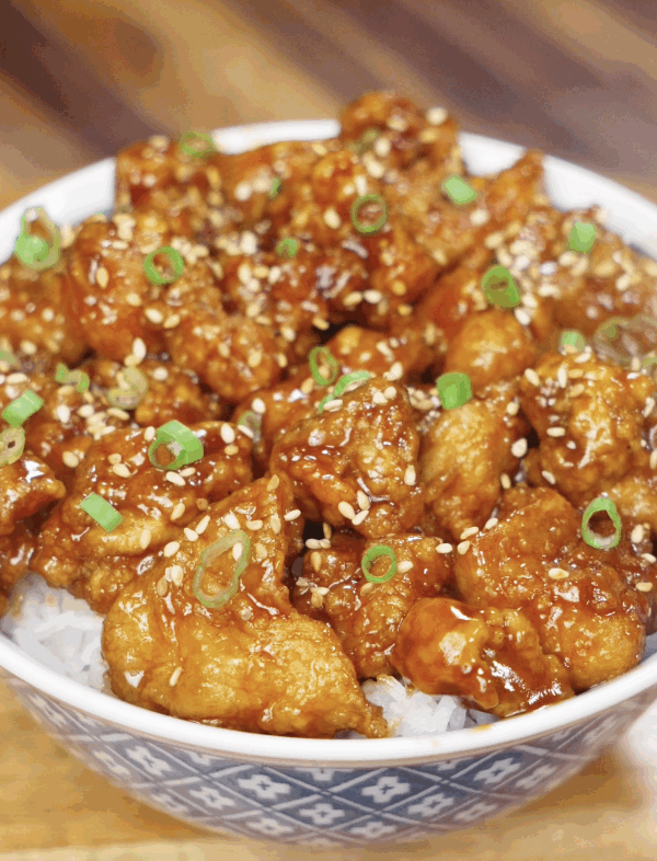 A bowl of General Tso’s Chicken served over white rice, topped with sesame seeds and sliced scallions.