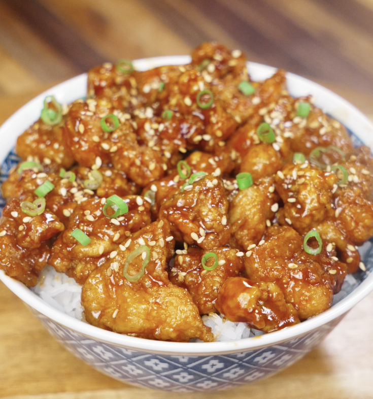 A bowl of General Tso’s Chicken served over white rice, topped with sesame seeds and sliced scallions.