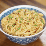 A bowl of peanut butter noodles sits on the table, topped with scallions, sesame seeds, and chili flakes for extra flavor.