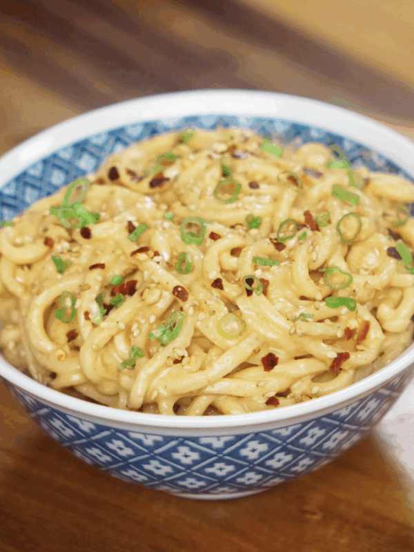 A bowl of peanut butter noodles sits on the table, topped with scallions, sesame seeds, and chili flakes for extra flavor.