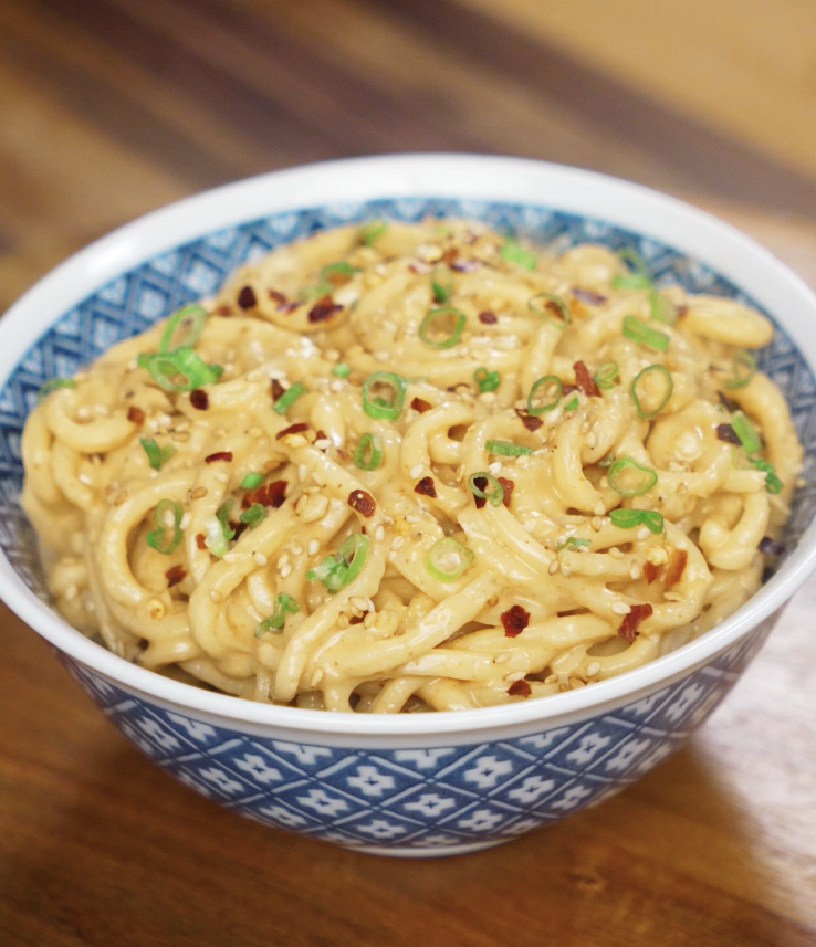 A bowl of peanut butter noodles sits on the table, topped with scallions, sesame seeds, and chili flakes for extra flavor.