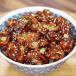 A full bowl of crispy sesame chicken coated in a thick, shiny glaze and topped with scallions and sesame seeds.